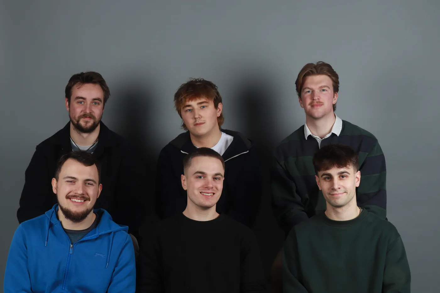 Six members of the film's head crew in a studio portrait, three standing behind three seated, against a grey backdrop